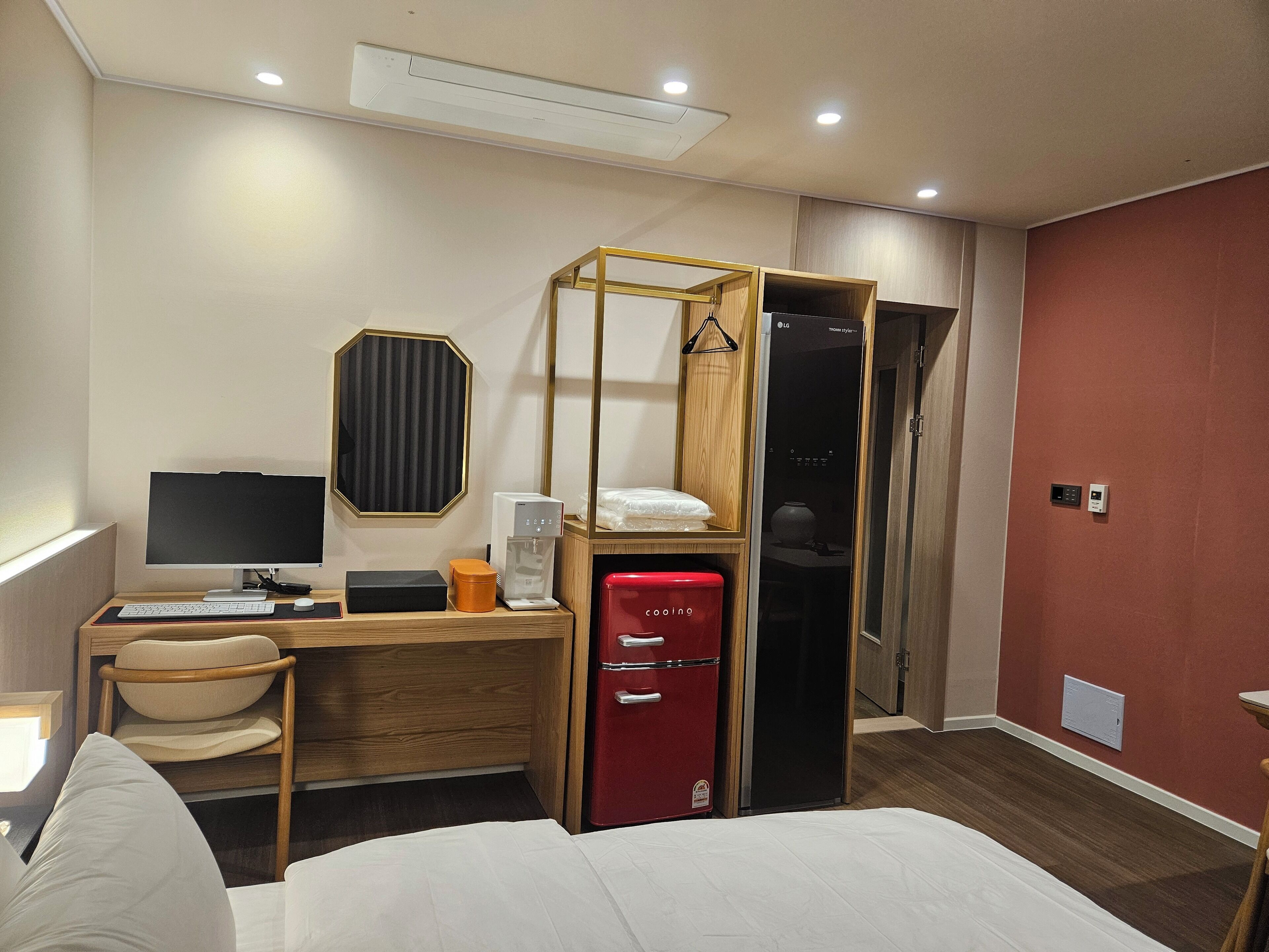 Una habitación de hotel con una cama, un escritorio con computadora, un refrigerador rojo, un armario de madera y un espejo.