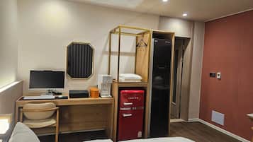 Una habitación de hotel con una cama, un escritorio con computadora, un refrigerador rojo, un armario de madera y un espejo.