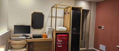 A hotel room with a bed, a desk with a computer, a red refrigerator, a wooden wardrobe, and a mirror.