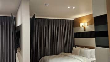 A hotel room with a bed, curtains, a wall-mounted TV, and a ceiling vent.