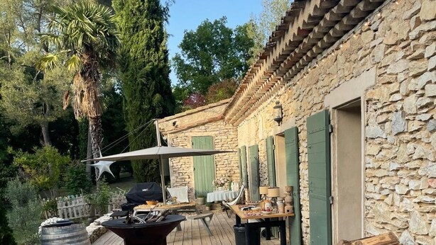 Eine Holzterrasse mit Gartenmöbeln, einem Grill und einem Steinhaus mit grünen Fensterläden.