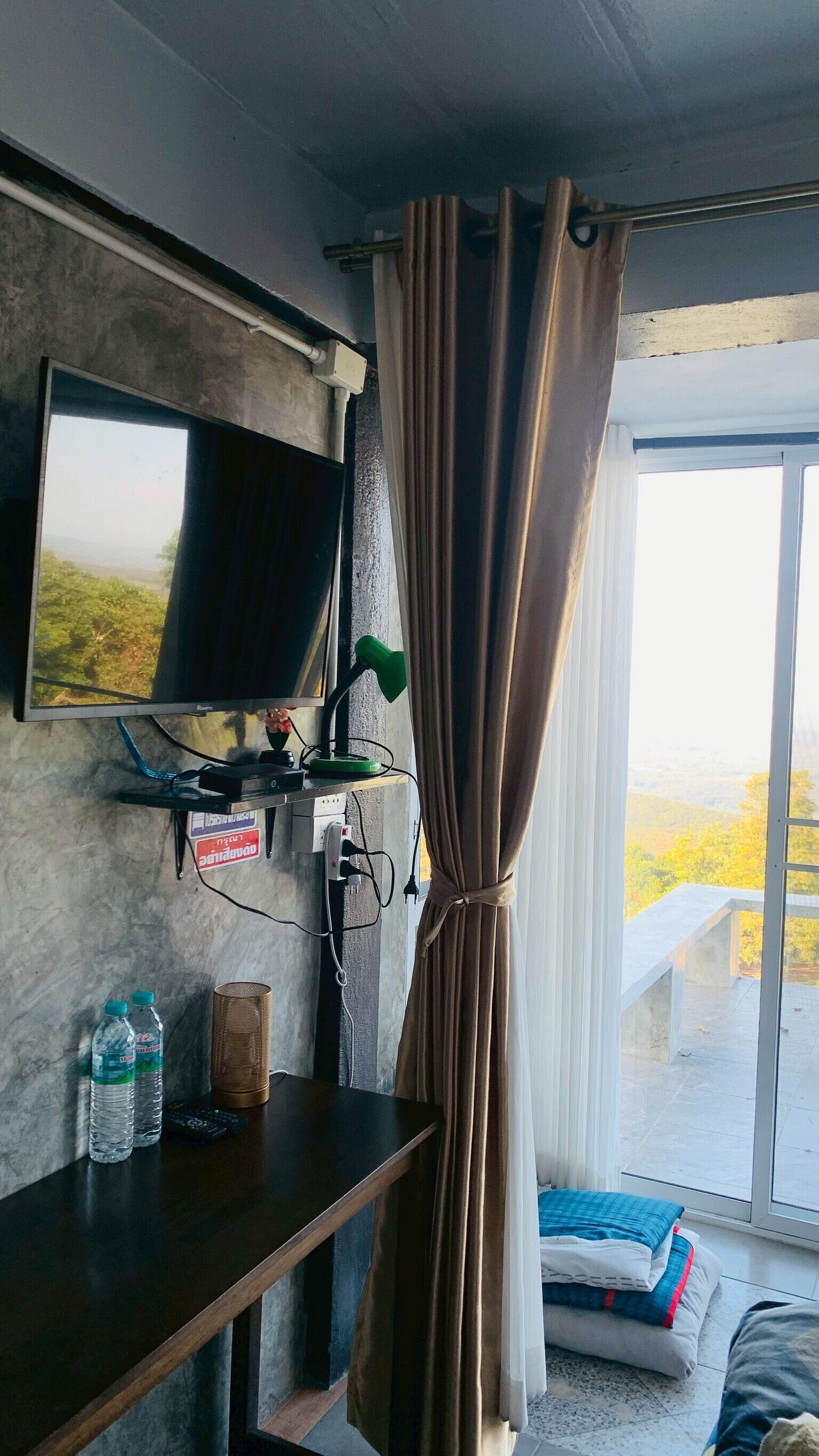 A room with a TV mounted on the wall, a desk with a lamp, two water bottles, and a bed with pillows.