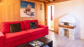 Un canapé rouge agrémenté de coussins à motifs, un plafond en bois avec des poutres apparentes, une cheminée en pierre et un tableau encadré accroché au mur.