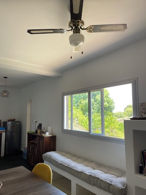 A room with a ceiling fan, a window with a view of greenery, a bench, and a dining table with a chair.
