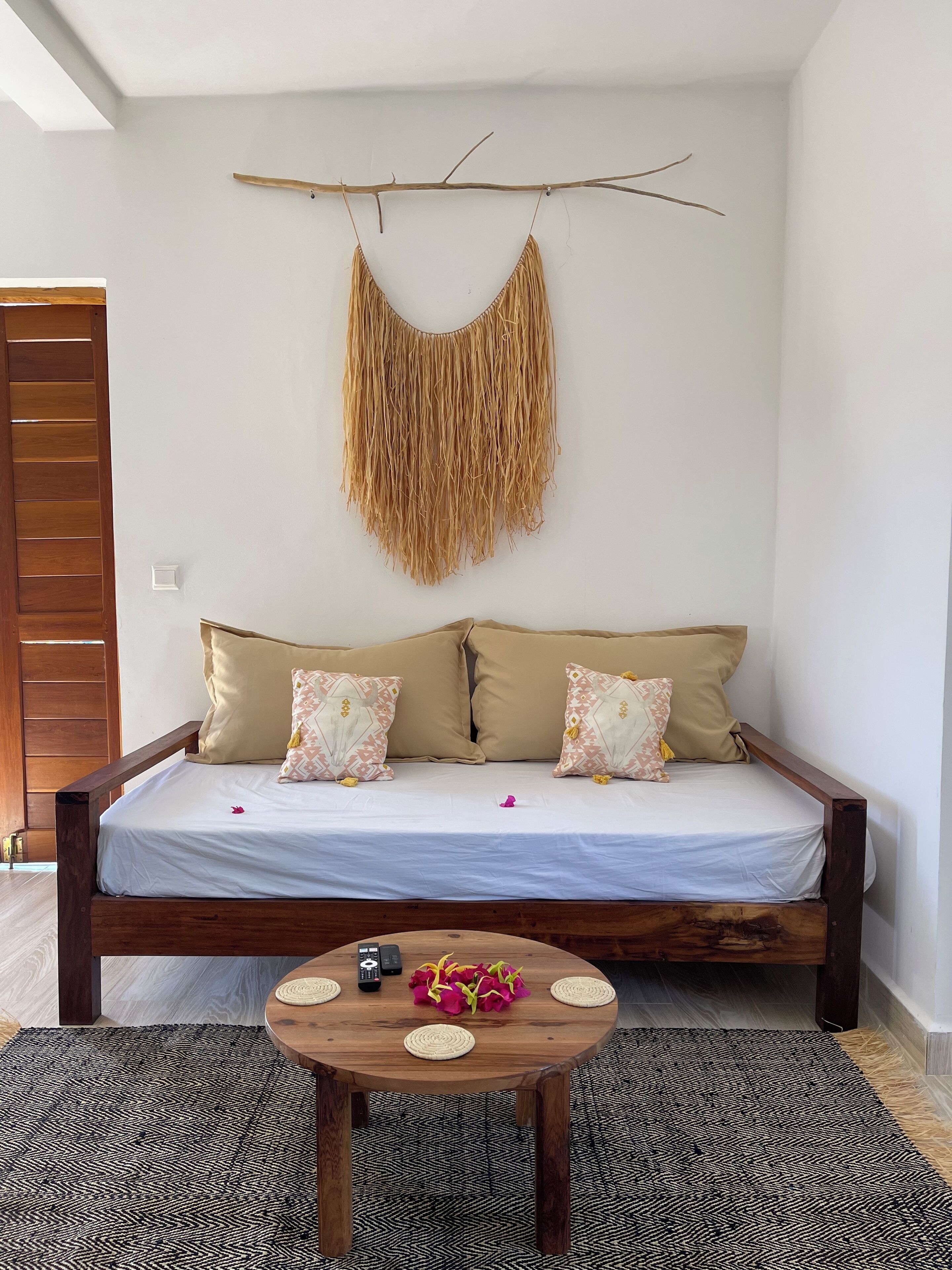 A room with a wooden bed, two patterned pillows, a woven wall hanging, and a round wooden table with a woven mat.