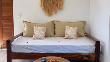 Une chambre avec un lit en bois, deux coussins à motifs, une décoration murale tissée et une table ronde en bois surmontée d’un tapis tissé.