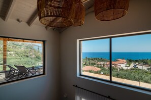 Ein Zimmer mit zwei großen Fenstern, Weidenkorb-Deckenleuchten und Blick auf das Meer und Grünflächen.