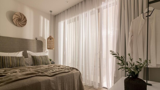 A bedroom with a bed, curtains, a bedside table, a mirror, and a wall decoration. - Cossoro Cave Villa Vasilikos (Vasilikos)