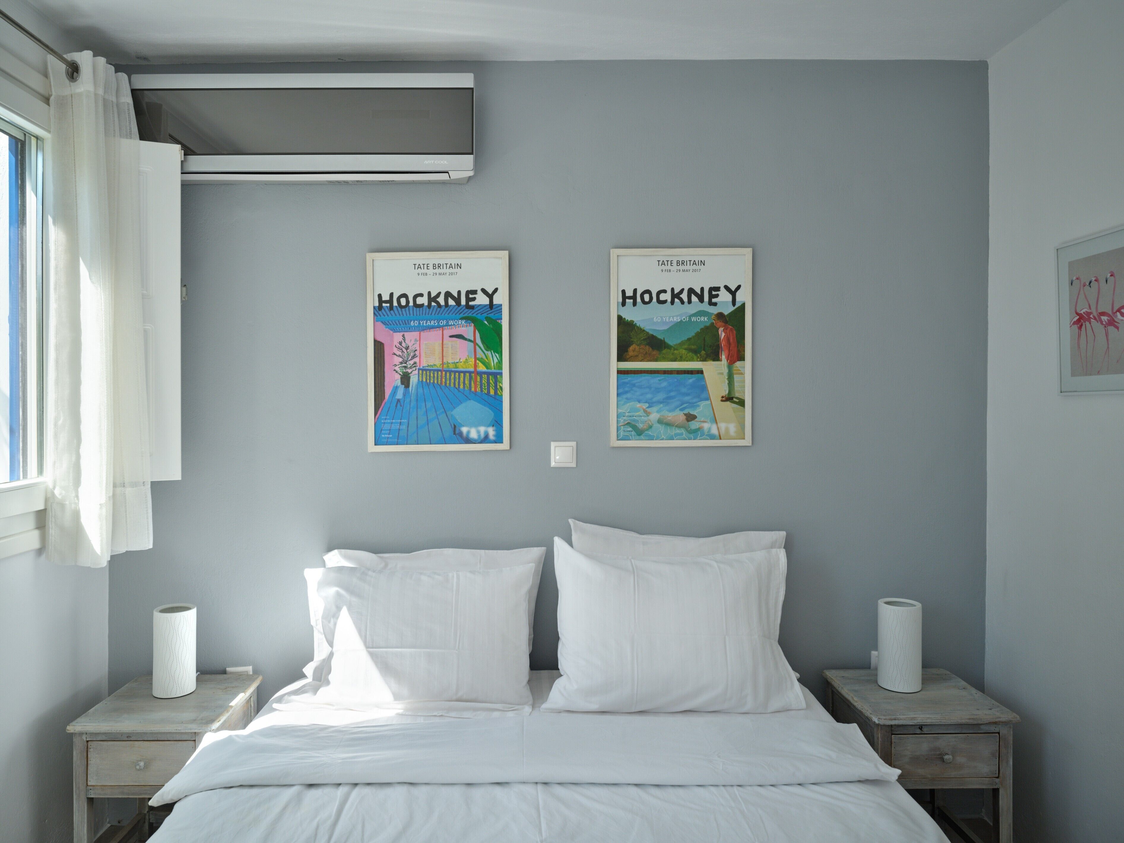 A bedroom with a bed, two nightstands, a window with curtains, and two framed posters on the wall.