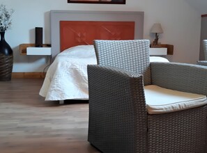 A room with a bed, a grey wicker chair with a cushion, a bedside table with a lamp, and a vase with dried flowers.