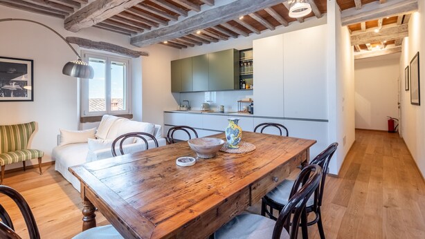 Een eethoek met een houten tafel, stoelen, een vaas en een keuken op de achtergrond.