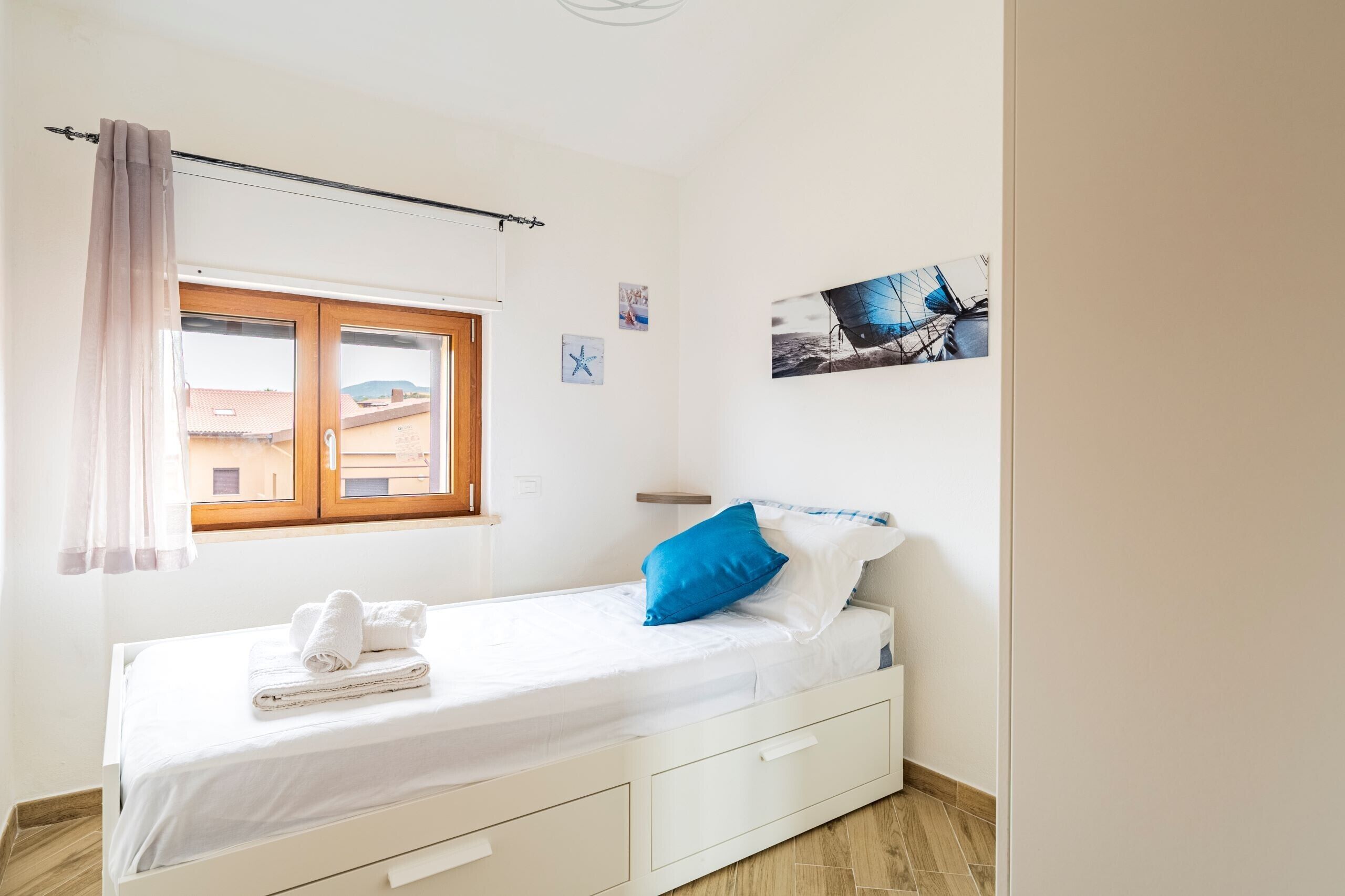 Un lit simple avec un matelas blanc, un oreiller bleu et une couverture blanche. Il y a une fenêtre avec un rideau, un tableau accroché au mur et une petite étagère.