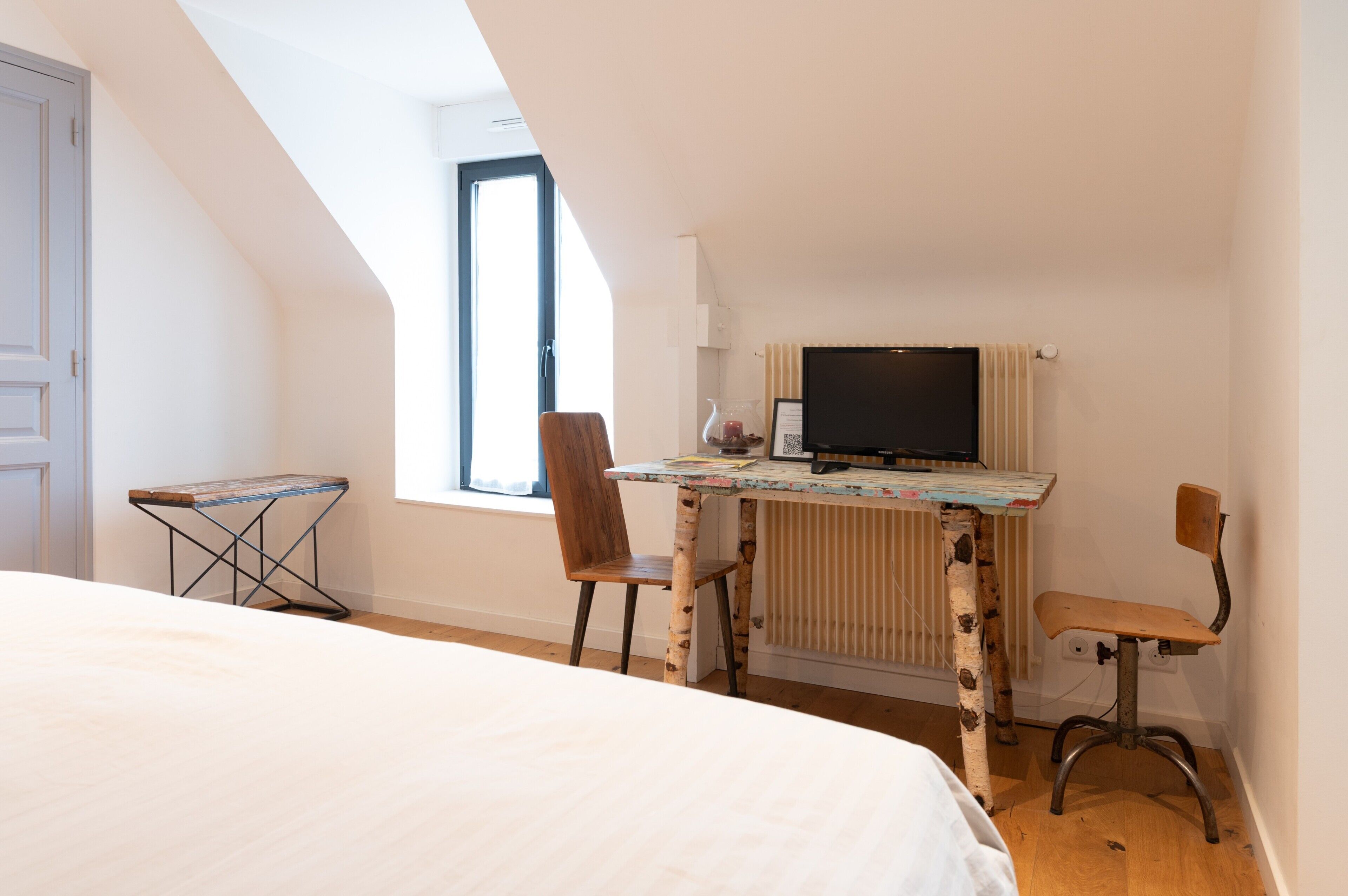 A room with a bed, a desk with a computer, two chairs, a radiator, and a window.