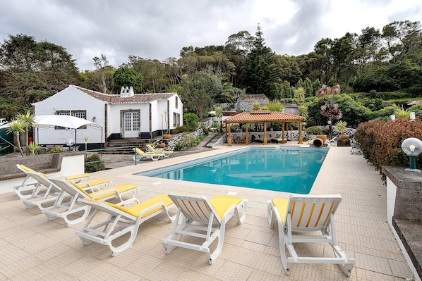 Una piscina con sillas de descanso, un pabellón y un edificio blanco con tejado de azulejos.