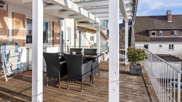A covered outdoor deck with a table and chairs, a potted plant, and a view of a building with a tiled roof. - Apartment 'Terrassenblick' with Mountain View, Private Terrace, and Wi-Fi (Sankt Georgen)