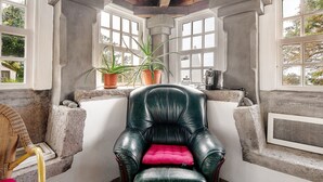 A corner room with a black leather chair, a wooden chair, and a pink cushion. There are potted plants on a ledge and a window with a view of trees. - Double Room 3 – Quinta do Rossio - Rural Tourism (Biscoitos)
