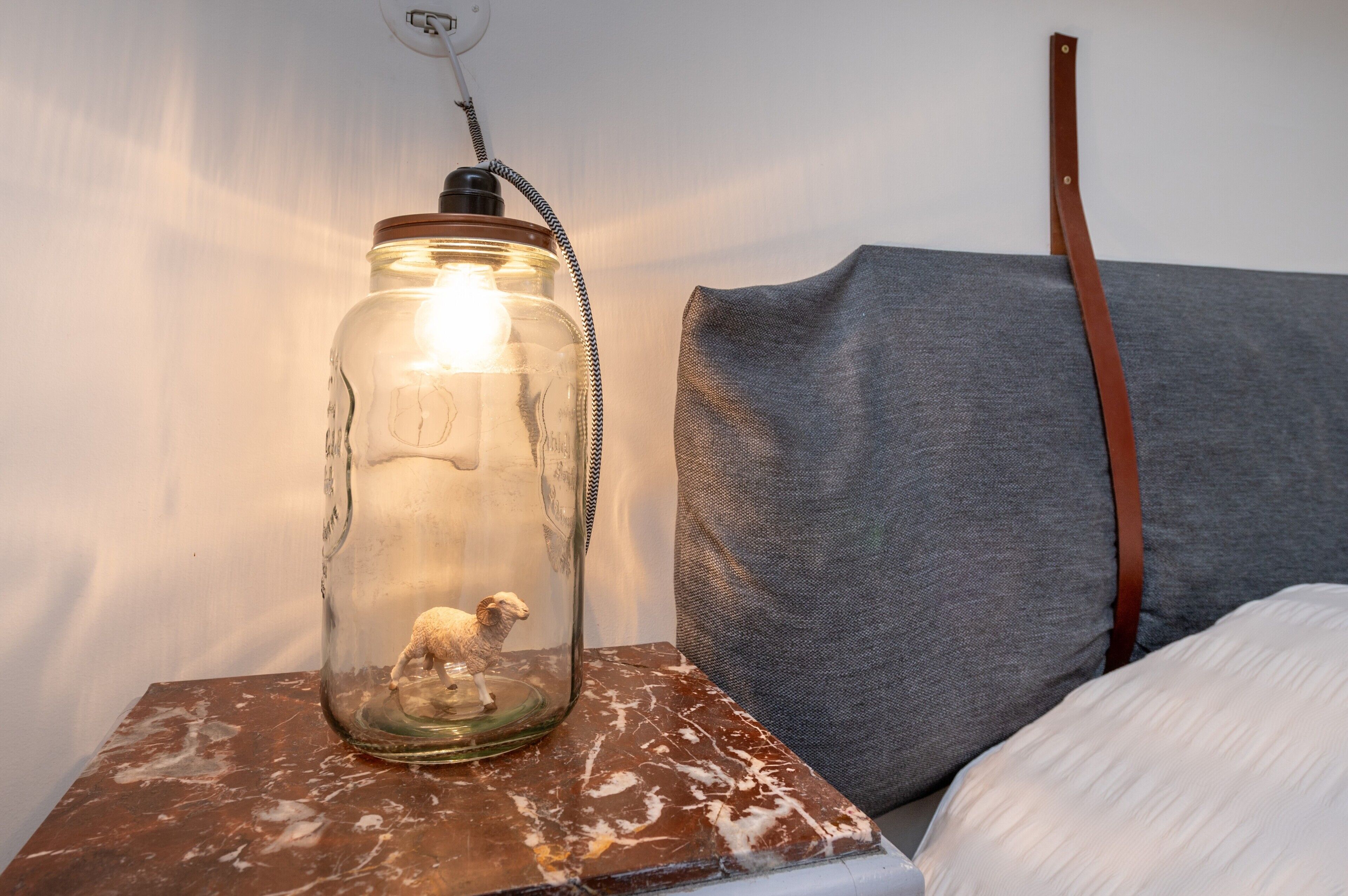 Une lampe de style rustique, en forme de bocal, ornée d’une figurine de mouton, posée sur une surface en bois, à côté d’un canapé gris recouvert de tissu.