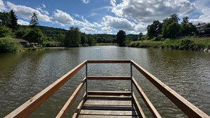 Un muelle de madera se adentra en un río tranquilo, con un paisaje forestal y un cielo parcialmente nublado al fondo.