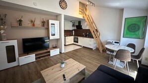 Un apartamento moderno con sala de estar, cocina y comedor, que incluye una escalera de madera, televisión empotrada y un cuadro de arte con motivos vegetales.