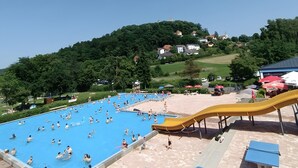 Una piscina con gente, un tobogán y casas en una colina al fondo.