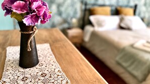 Ein Holztisch mit einer Spitzendecke, eine Vase mit violetten Blumen und ein Bett mit Kissen im Hintergrund.