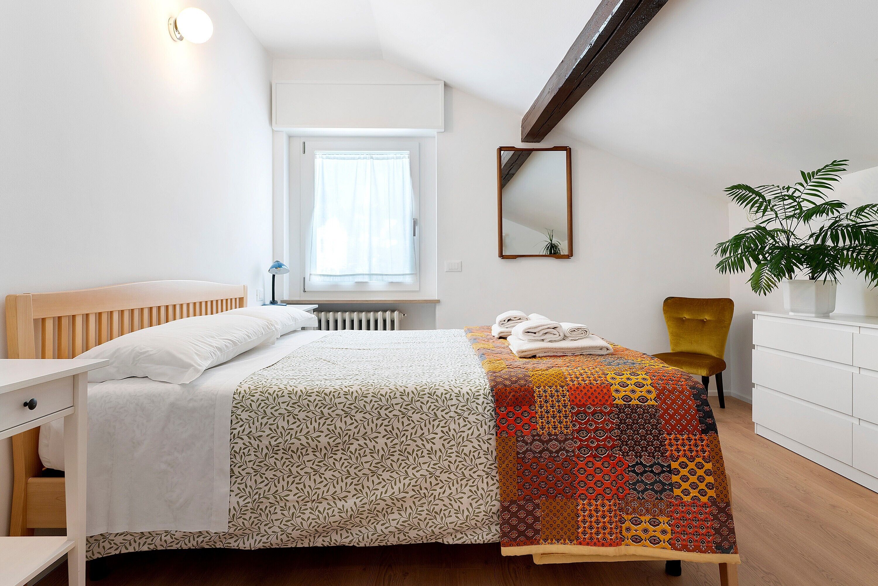 Une chambre à coucher avec un lit, une chaise, une commode, une fenêtre avec des rideaux, un miroir et une plante.