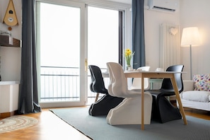 Ein modernes Zimmer mit Esstisch, Stühlen, einer Couch, einer Lampe und Blick durch ein großes Fenster.