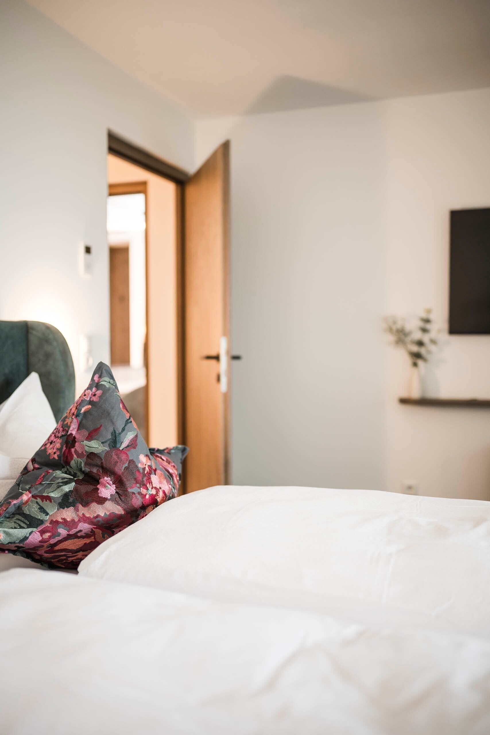 A bed with white bedding and a floral pillow, a wooden door, and a small shelf with a plant.
