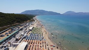 Une plage bordée de rangées de parasols, un parking et des montagnes en arrière-plan.