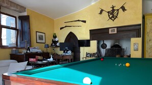 Una mesa de billar en una habitación con paredes amarillas, una chimenea y armas colgadas.