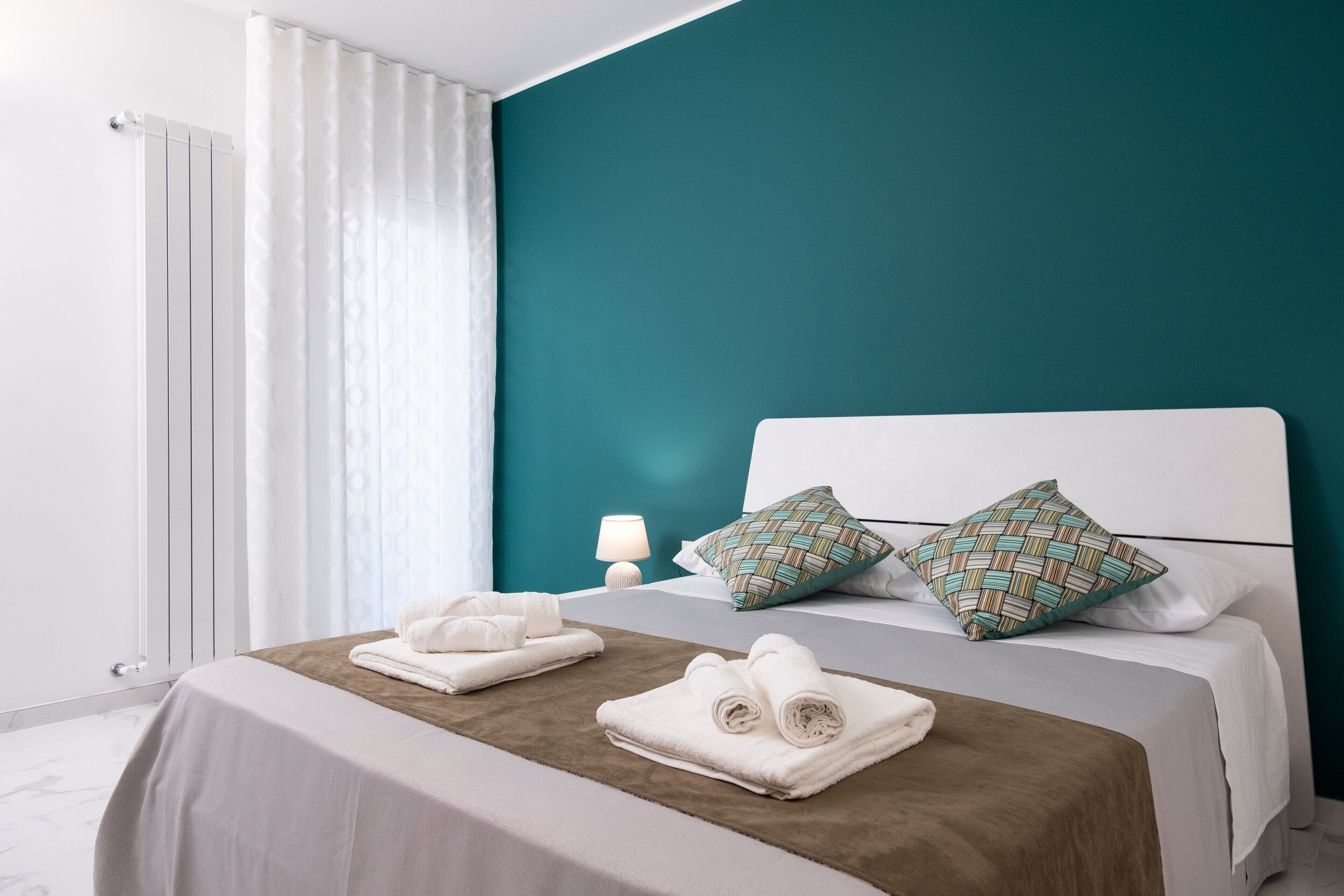 Una cama bien hecha con toallas, una lámpara de noche y almohadones estampados en una habitación con paredes color turquesa y ropa de cama blanca.