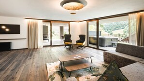 Ein modernes Wohnzimmer mit Flachbildfernseher, einer Couch, zwei Sesseln, einem Couchtisch und einer großen Schiebeglastür, die auf einen Balkon mit Whirlpool führt.