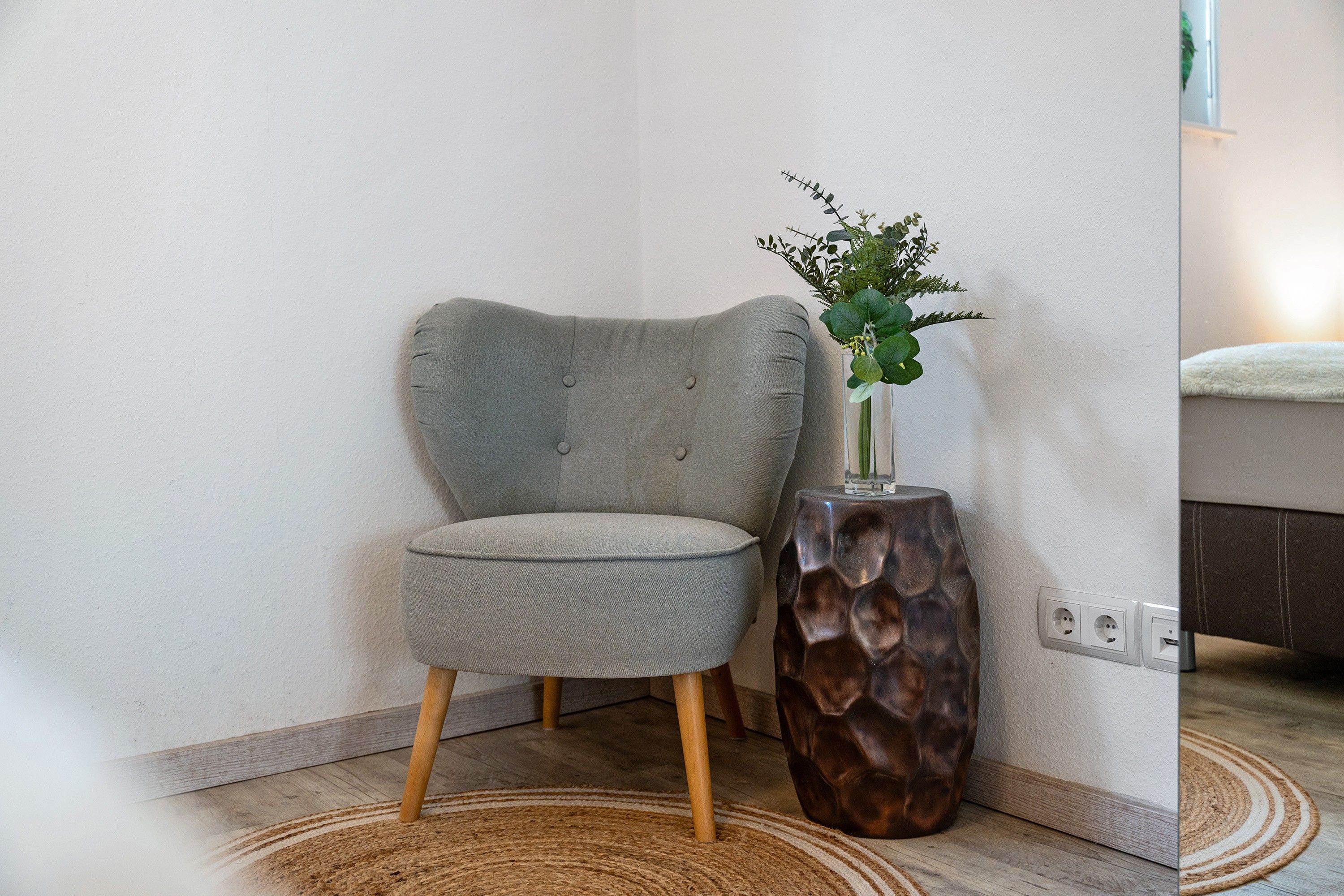 Ein Teil eines Zimmers mit einem grau gepolsterten Sessel, einem Holznachttisch mit einer Vase voller Grünpflanzen und einem gewebten Teppich auf dem Boden.