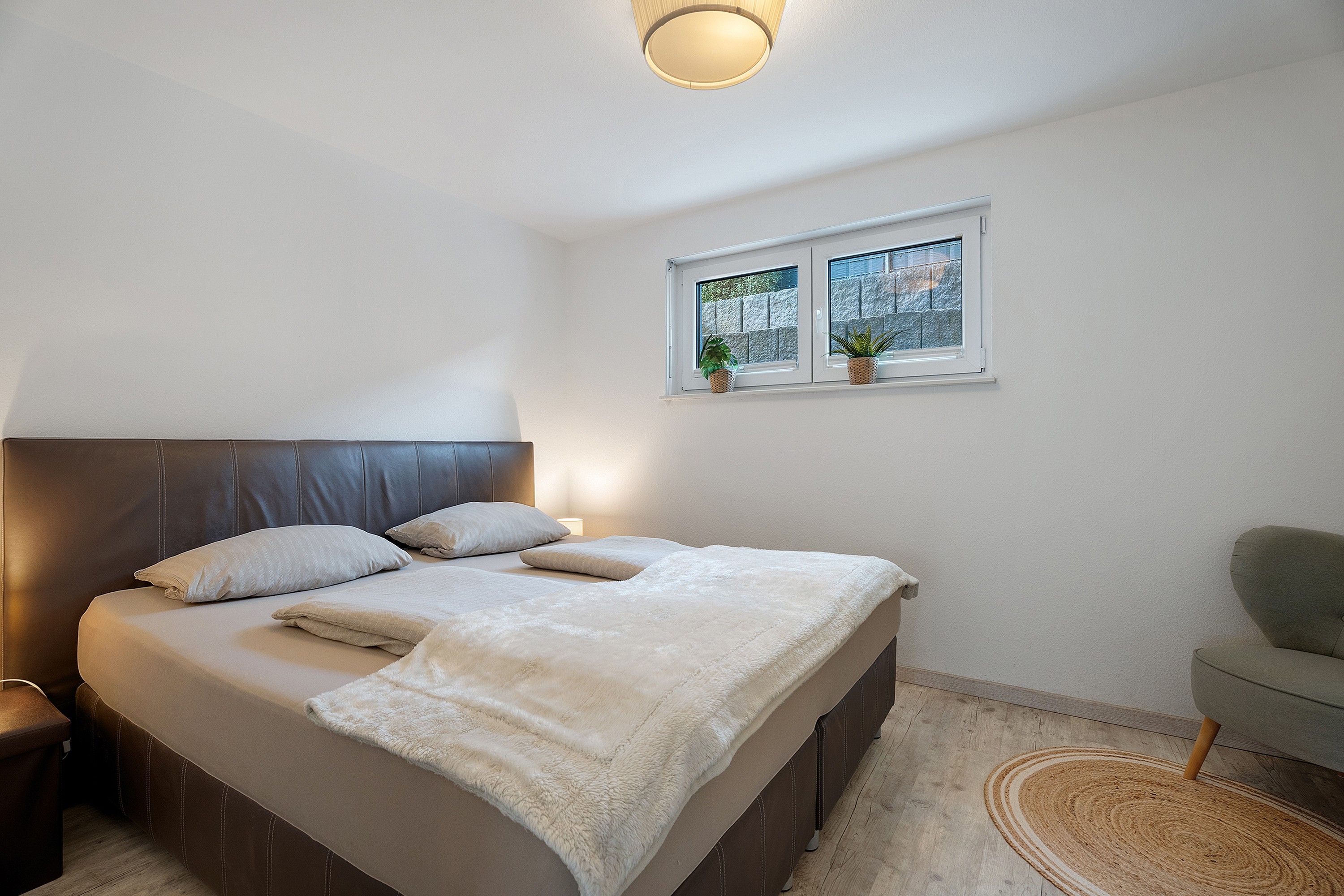 Ein Schlafzimmer mit einem Bett, Kissen, einem Nachttisch, einer Lampe, einem Fenster mit Pflanzen und einem Sessel.