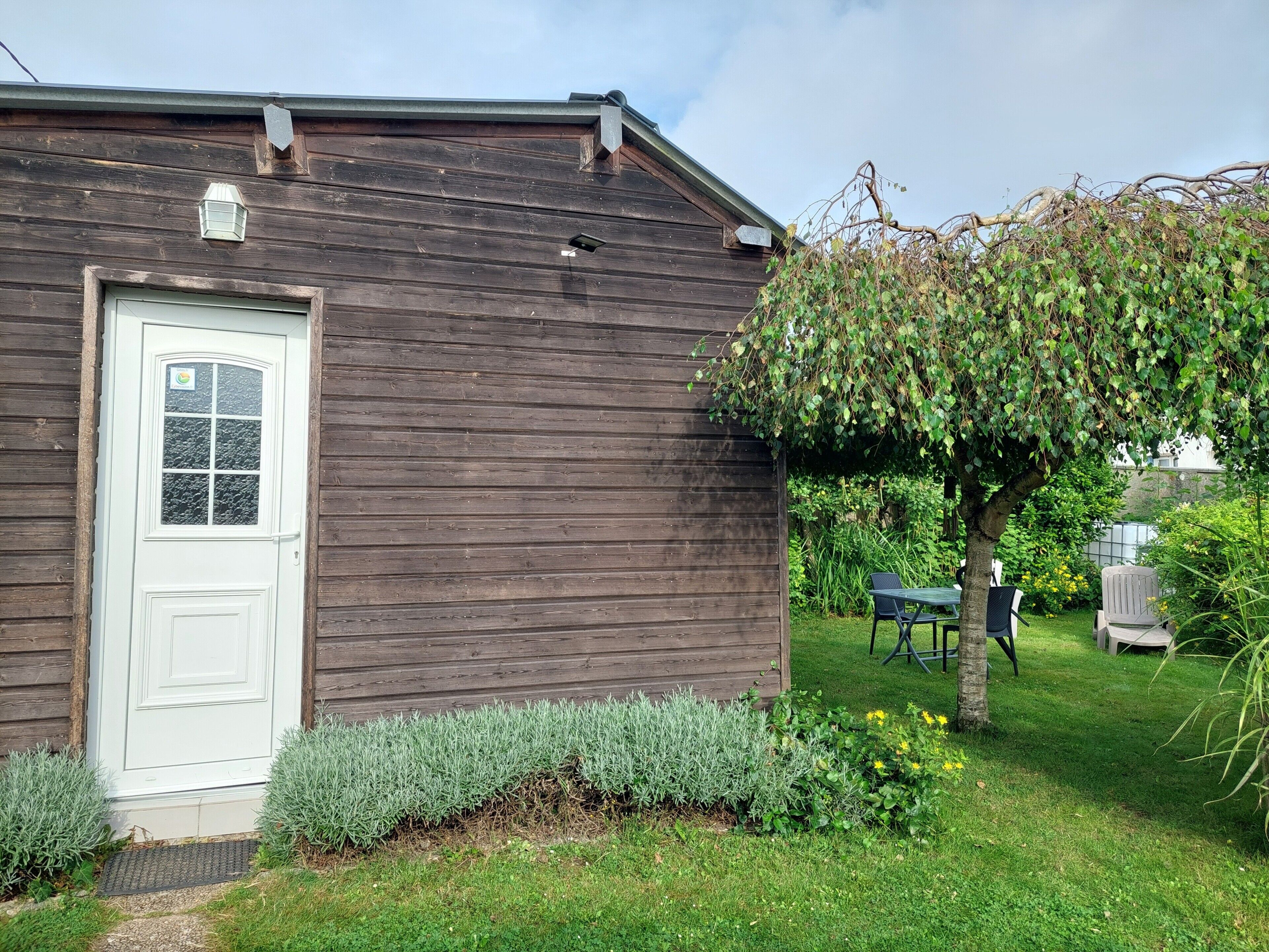 Un bâtiment en bois avec une porte et une fenêtre blanches, entouré d’un jardin avec de la pelouse, des arbustes et un arbre.
