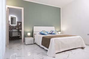 A bedroom with a bed, bedside tables, a nightstand, a mirror, and a bathroom with a sink and patterned tiles. - Guest House 'Verde Salvia Prestige' with Shared Terrace, Wi-Fi and Air Conditioning (Marsala)