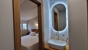A bedroom with a bed, a nightstand, a bedside table, a mirror, and a bathroom with a sink.