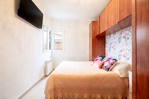 A bedroom with a bed, a TV mounted on the wall, a radiator, wooden cabinets, and a window with curtains.
