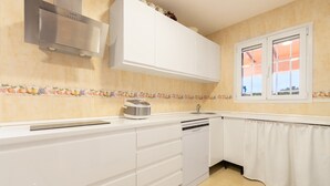 Une cuisine avec des armoires blanches, une hotte de cuisinière en acier inoxydable, un lave-vaisselle, une fenêtre avec des rideaux et un dosseret carrelé orné de motifs de fruits.
