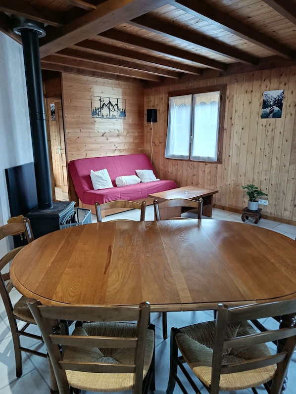 A wooden interior with a round wooden table surrounded by chairs, a red sofa, a black heater, and a window with curtains.