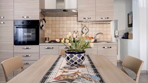 A modern kitchen with light wood cabinets, a black oven, and a decorative vase with flowers on the dining table. - Casa Jessica – Your Stay Between the Sea, Relaxation, and Authentic Sicily (Balestrate)