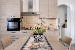 A modern kitchen with light wood cabinets, a black oven, and a decorative vase with flowers on the dining table. - Casa Jessica – Your Stay Between the Sea, Relaxation, and Authentic Sicily (Balestrate)