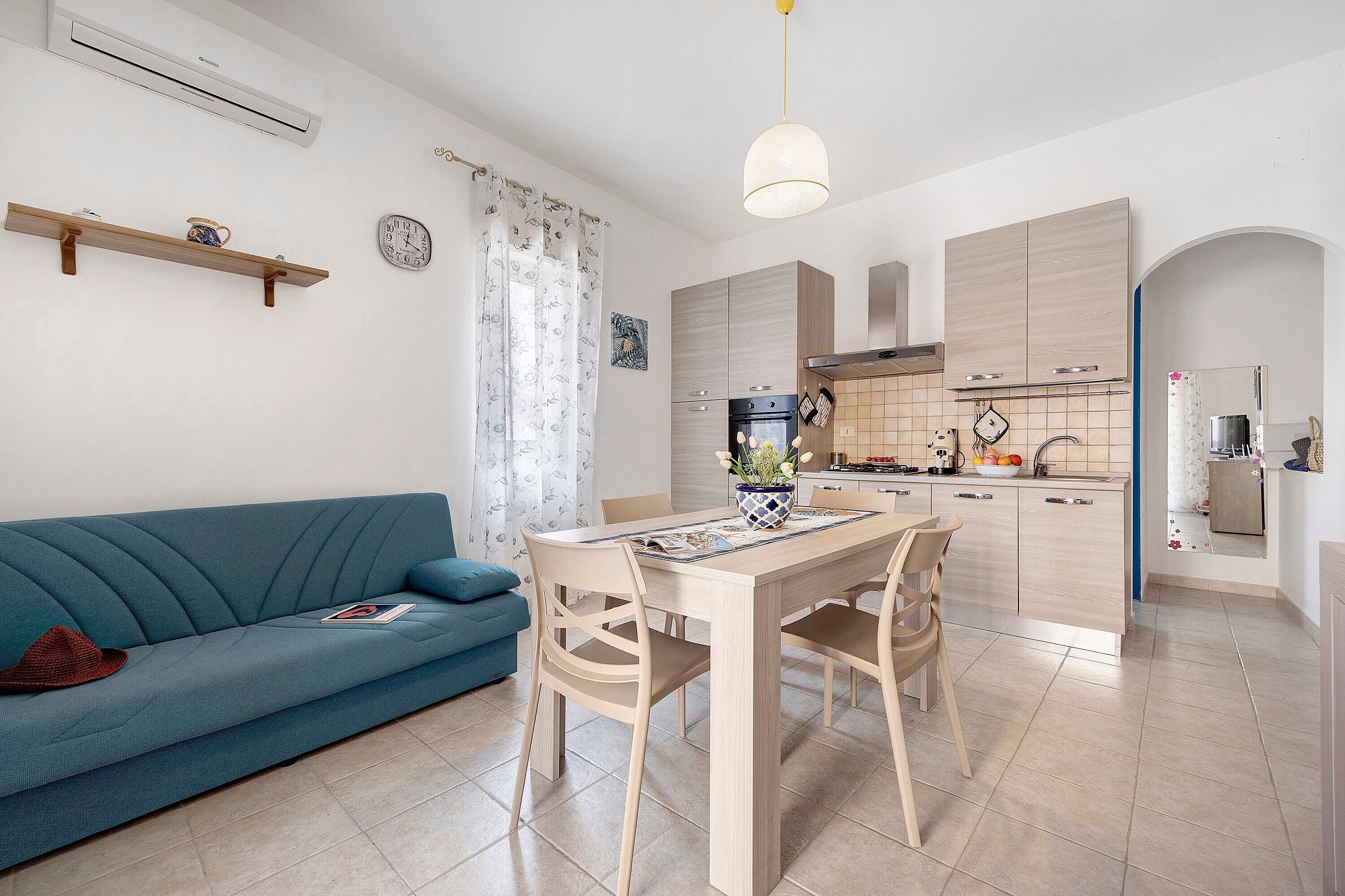A modern kitchen with wooden cabinets, a dining table, and chairs. There is a blue sofa with a hat on it.