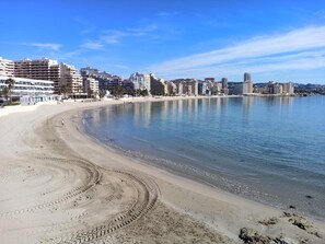 A beach with a curved shoreline, buildings in the background, and a clear sky.
