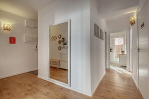 A hallway with a wooden floor, a mirror, and a bench. There are wall-mounted lights and a door leading to another room.