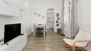 A modern living room with a flat-screen TV, a dining table with chairs, a white armchair, and a kitchen area in the background.