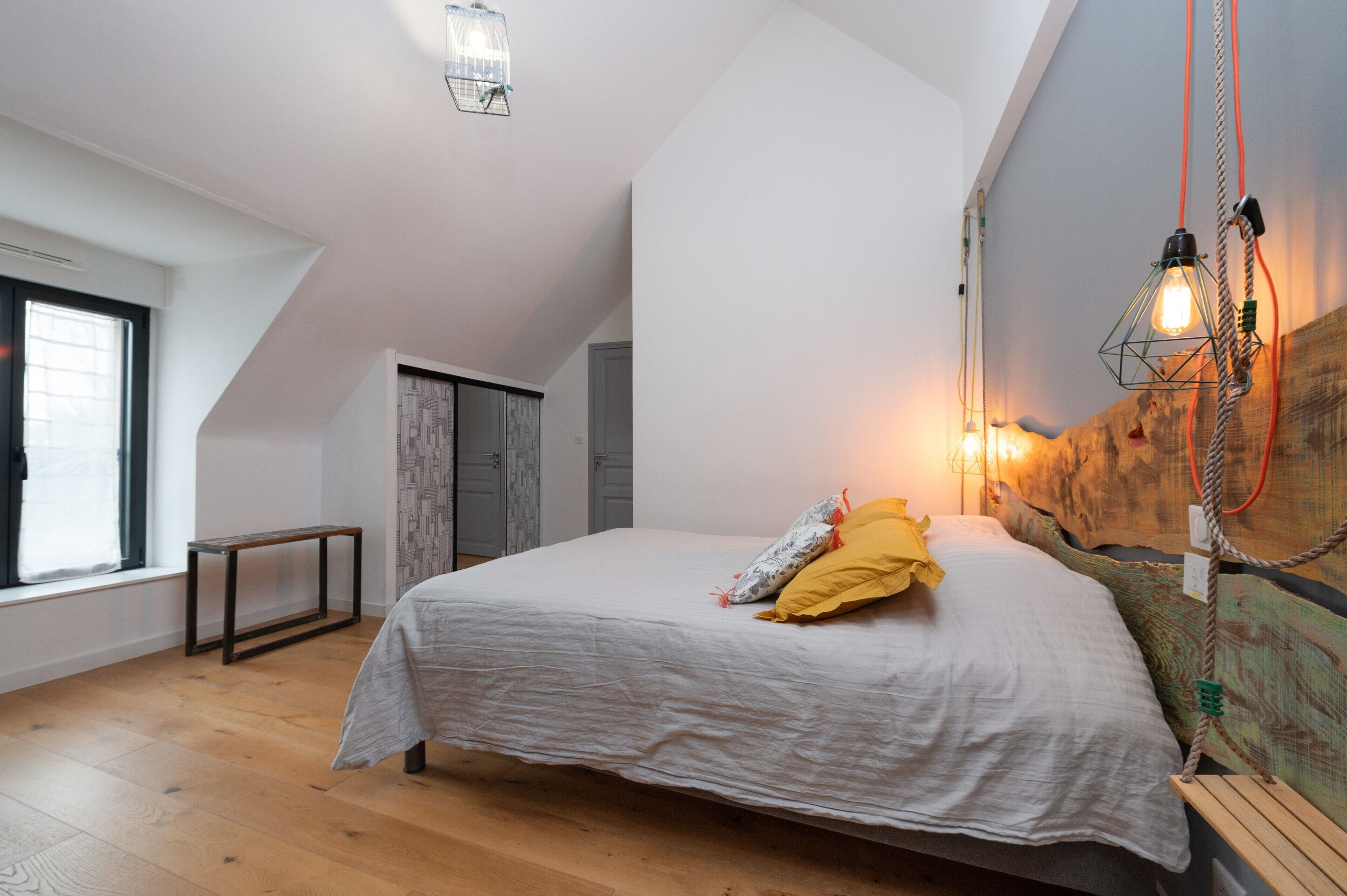 Un dormitorio con techo inclinado, una cama con ropa de cama blanca, un banco de madera, una mesita y un aplique de pared.