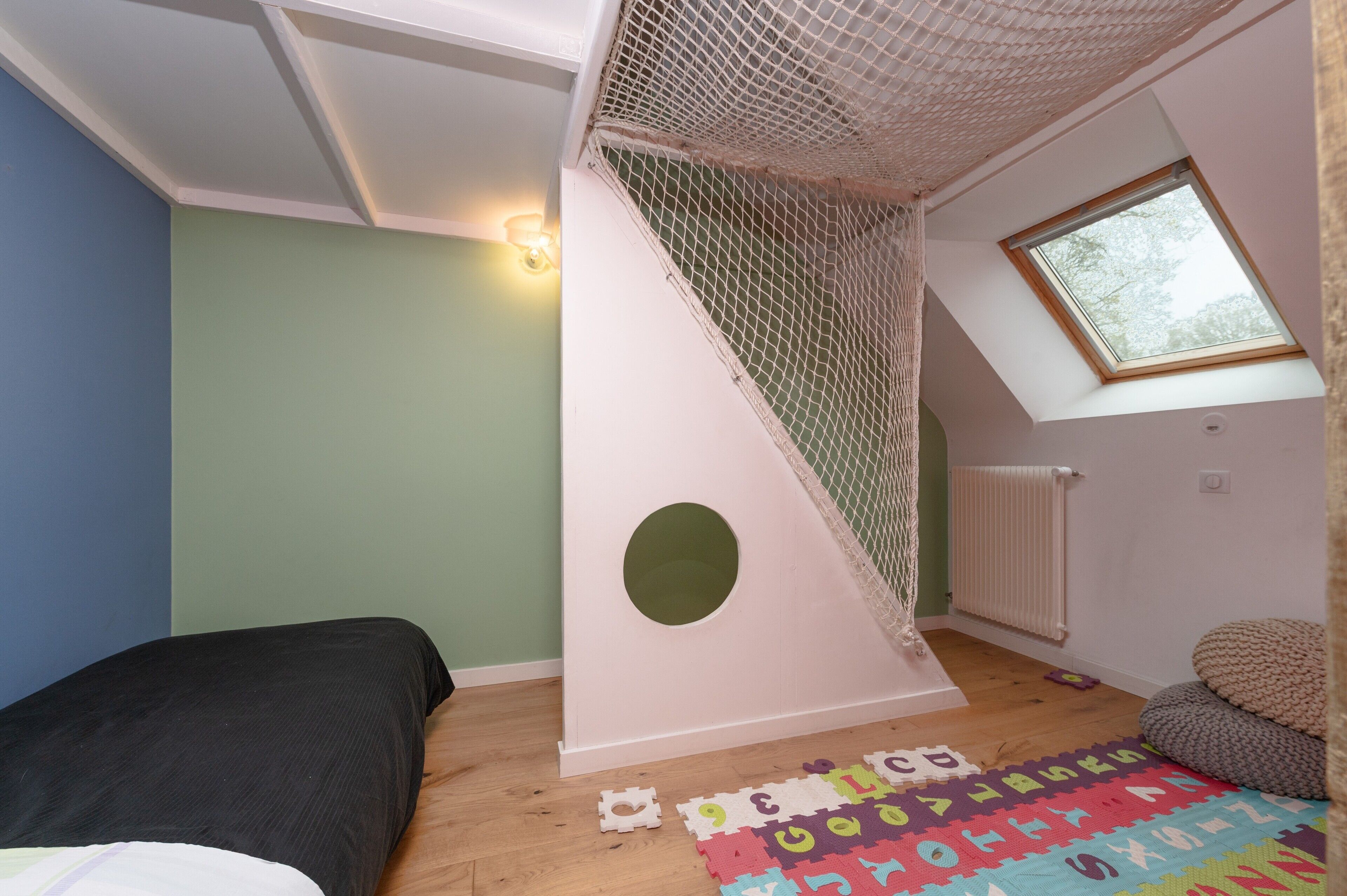 A room with a sloped ceiling, a window, a bed, a play area with a net, and a radiator.