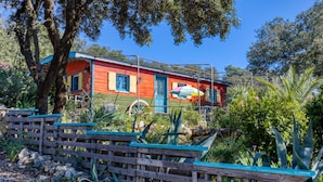Une maison rouge avec une porte et des fenêtres bleues, entourée d’arbres et de plantes.