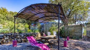 Un espace extérieur aménagé avec une zone de repos couverte, comprenant une table et des chaises, un fauteuil et une structure en forme de pergola.